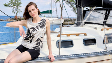 Women's sleeveless linen tank top with black tropical palm leaf print on natural beige background, styled with black biker shorts, worn on a sailboat by the water.