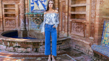 Woman wearing a floral off-shoulder blouse and cropped wide-leg jeans standing by a Spanish tiled fountain.
