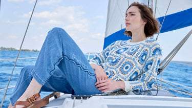 A woman wearing a blue and white geometric crochet sweater with wide leg denim pants, reclining on a sailboat.