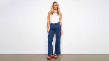 A woman wears dark indigo denim jeans in a wide-leg cut, styled with a white tank top and brown platform sandals.