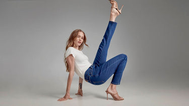 Woman in a floor pose wearing skinny indigo jeans and a white top, one leg extended overhead in heeled sandals.