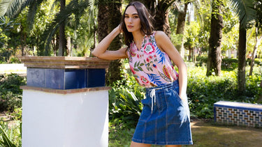 A woman in a Charlie B sleeveless pink floral print top with a denim drawstring mini skirt, posing in a lush tropical garden.