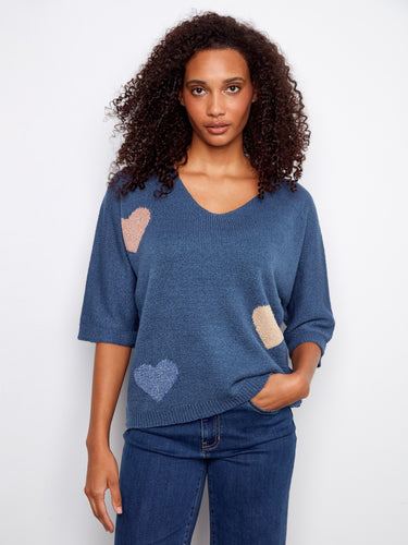 Blue sweater with a V-neckline featuring heart patterns, crafted for a relaxed fit. The elbow sleeves add a casual touch. By Charlie B.