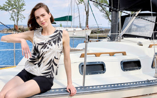 Women's sleeveless linen tank top with black tropical palm leaf print on natural beige background, styled with black biker shorts, worn on a sailboat by the water.