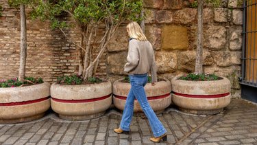 A woman walking beside planted stone flower beds, wearing a beige sweater and jeans flared at the bottom, in front of an old stone wall.