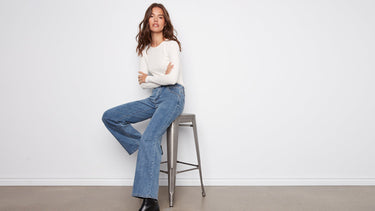 Woman sitting on a stool wearing a white sweater and blue wide leg jeans against a plain background