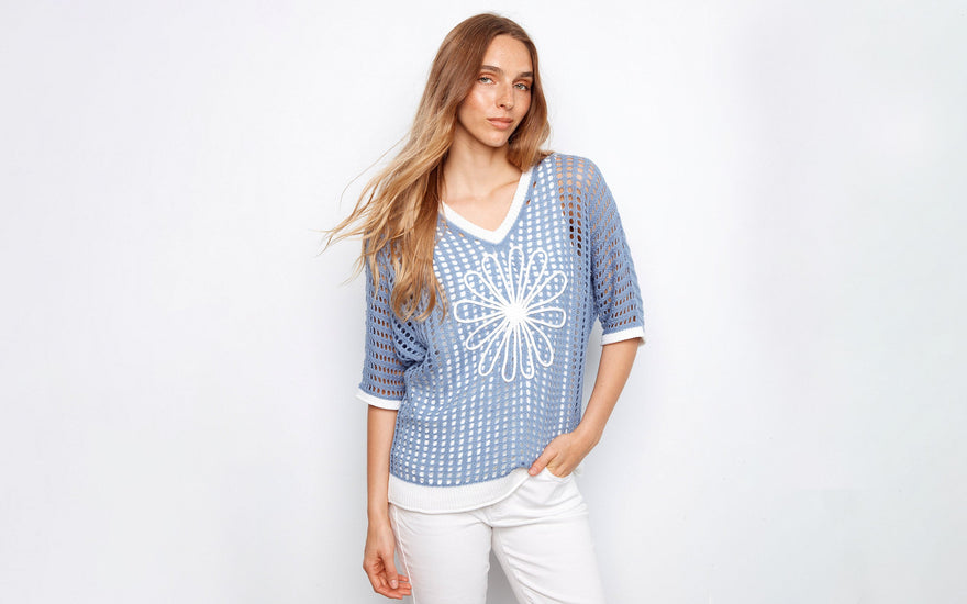 A woman wearing a blue open-knit sweater with a white flower design and white pants.