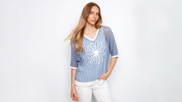 A woman wearing a blue open-knit sweater with a white flower design and white pants.
