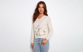 A woman wearing a beige V-neck sweater with floral embroidery and light wash denim jeans.