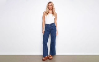 A woman wears dark indigo denim jeans in a wide-leg cut, styled with a white tank top and brown platform sandals.