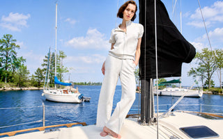 A woman wearing a light grey zip-neck top and matching wide-leg pants, standing on a sailboat near a marina.