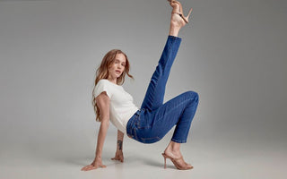 Woman in a floor pose wearing skinny indigo jeans and a white top, one leg extended overhead in heeled sandals.