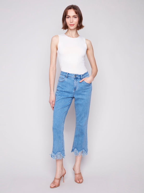 Woman in sleeveless white top and cropped light blue jeans with fringed hems, posing in nude high-heel sandals against neutral background