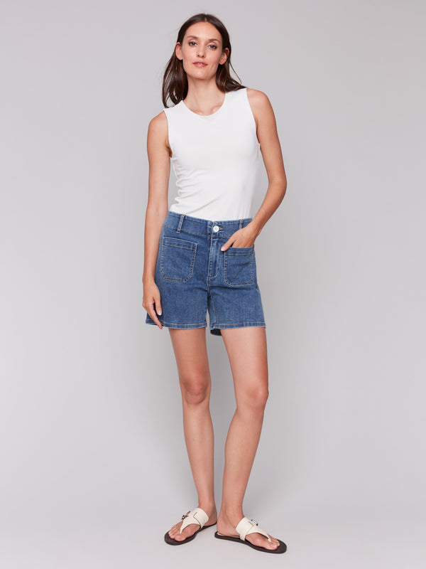 Woman in a sleeveless white top with blue denim shorts featuring front patch pockets, paired with white sandals, standing on grey background.