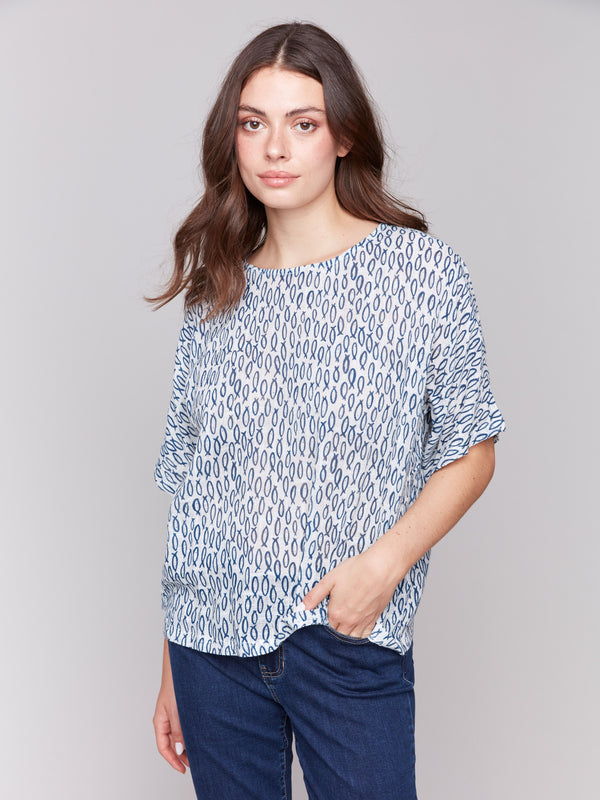 Woman wearing a loose short-sleeve blouse with a blue and white abstract oval print, paired with dark blue jeans, standing against a grey background