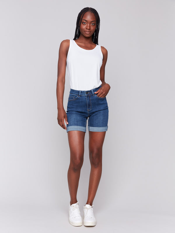 Woman wearing a sleeveless white top and dark blue rolled-hem denim shorts, paired with white sneakers, standing casually.