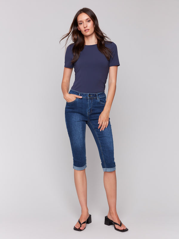 Woman wearing a fitted short-sleeve navy top with dark blue cuffed denim capri pants and black heeled sandals, standing against a plain grey background.