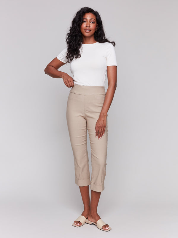 Woman wearing a fitted white short-sleeve top with beige cropped pants featuring cuffed hems, paired with beige slide sandals, on grey background.